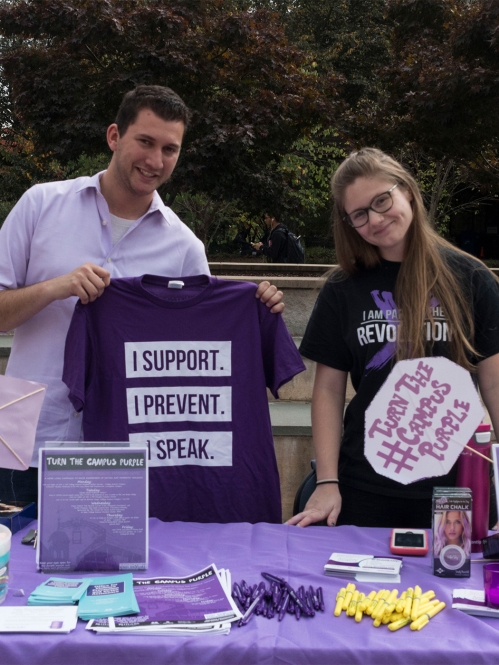two students work stand behind a table with flyers, t-shirts, and giveaways to promote Turn the Campus Purple on Livginston camppus